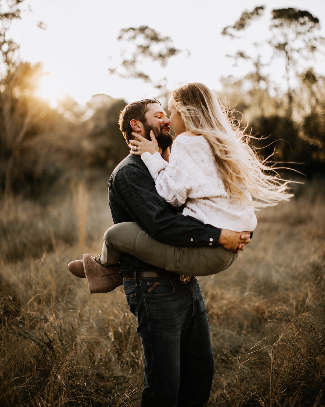 Un homme enlace tendrement sa compagne dans un champ au coucher du soleil, illustrant la complicité et l'amour durable au sein d'un couple.