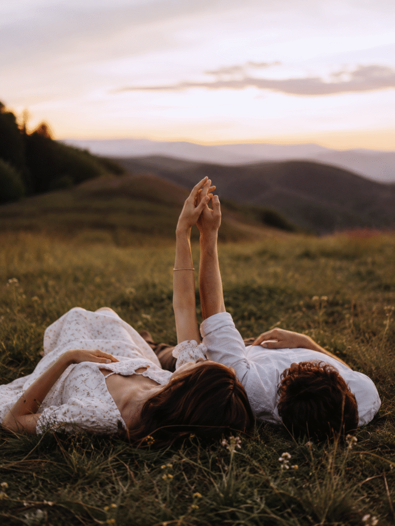 Un couple amoureux allongé dans une prairie au coucher du soleil, les mains enlacées vers le ciel, partageant un moment de complicité et de sérénité.